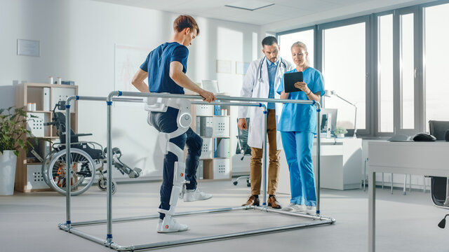 Modern Hospital Physical Therapy: Patient with Injury Walks Wearing Advanced Robotic Exoskeleton. Physiotherapy Rehabilitation Scientists, Engineers use Tablet Computer to Help Disabled Person