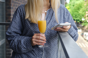 Fototapeta premium The girl holds a smartphone and drinks orange juice on the balcony. Fresh organic juice in a glass. Woman watching social networks, sending text messages or using apps on her mobile phone