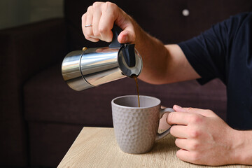 A man pours coffee from a stylish moka pot into a cup. Equipment for making coffee. Professional coffee brewing. Morning drink for breakfast. Aromatic black coffee in a cup close-up