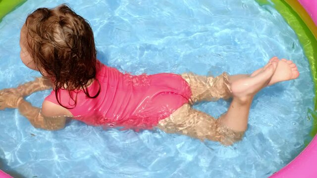 A girl in a swimsuit splashes in a small inflatable pool lying on her stomach and tries to swim. On a hot summer day, the baby is refreshed by swimming in the pool