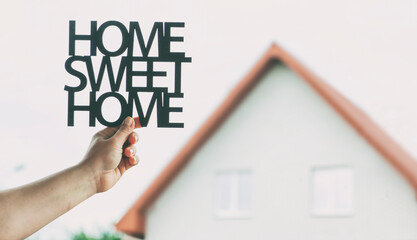 Hand holds wooden phrase Home sweet home in front of  blurred house. Concept of buying or building home of dreams