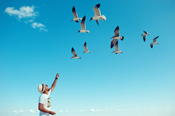 Naklejka premium Tourist man feeding flock of seagulls on the beach. Young male with straw hat, gives nachos to flying seabirds against clear blue amazing sky