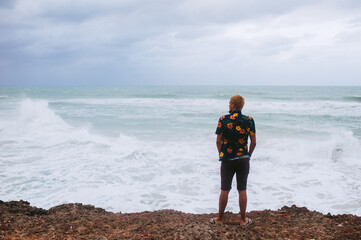 Back view of young male standing on the edge of the abyss facing to the sea with big waves, alone depressed person, power of nature, storm on seashore