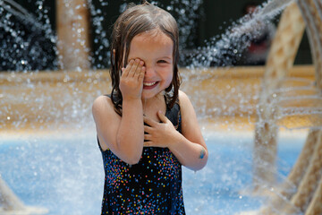 Happy little girl under fountains at a water park, wiping her eye