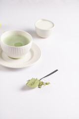 A white porcelain cup with japanese matcha tea drink on a white saucer plate on a white surface, a tea spoon with matcha powder, bamboos whisker