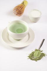 A white porcelain cup with japanese matcha tea drink on a white saucer plate on a white surface, a tea spoon with matcha powder, bamboos whisker