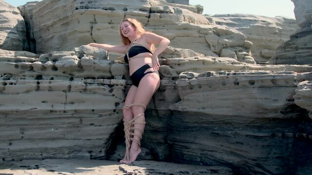 a dark-haired young girl, tied with ropes over her body and legs, stands by steep porous stones by the sea, a sunny day, her hair is loose