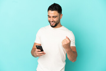 Young arab man isolated on blue background with phone in victory position