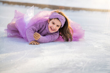 little girl in a pink sweater and a fluffy skirt on sunny winter day rides on open skating rink in...