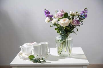 Obraz na płótnie Canvas Bouquet of hackelia velutina, purple and white roses, small tea roses, matthiola incana and blue iris in glass vase is on the white coffee table. Grey wall