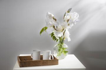 A bouquet of three white irises and a fern in a transparent vase on the table. Two ceramic tea in the tray. Breakfast
