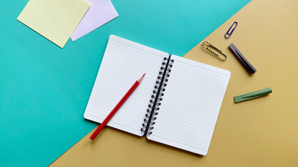 Top view flat lay of blank page notebook in a line with colored pencil, paper clips and pencil caps on green and yellow background