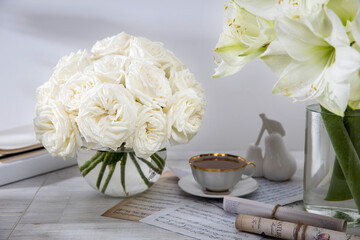 A bouquet of white roses in a round glass vase on a table with a cup of tea and a book. Copy space.