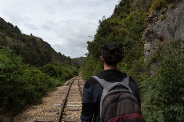 1 chico caucásico con pelo largo negro, maleta y saco negro caminando en las vías del tren rodeado por naturaleza 