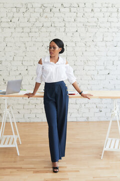 Business Black Woman Posing, Smiling. Successful Professional Concept. An African American Woman Stands In A Stylish Office In Front Of A Table And Looks To The Side