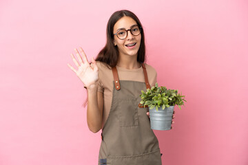 Young gardener girl holding a plant isolated on pink background saluting with hand with happy expression