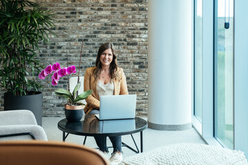 Portrait of Smiling Business Woman Working on a Laptop in  Office.

Beautiful casual businesswoman looking at camera while sitting in armchair with laptop computer and telecommuting from hotel lobby.