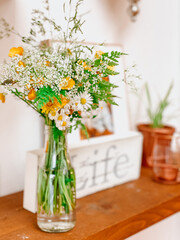 Wildflower vase glass bottle with daisies, dandelions, baby's breath, ferns on wooden scaffold shelf with copper bronze ornaments and life quote and family photo in background shelfie flowers