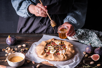 Process of making French galette with figs, goat cheese, pistachios and natural honey on baking paper on wooden table.  Woman in rustic closes adding honey.