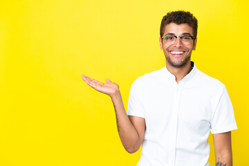 Young handsome Brazilian man isolated on yellow background holding copyspace imaginary on the palm...