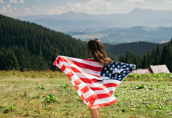 Back view young woman with american flag in hands standing on top of mountain. Female hiker waving national usa flag outdoor. Patriotic girl celebrating us independence day at vacation