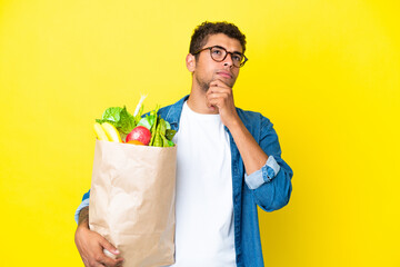 Young Brazilian man holding a grocery shopping bag isolated on yellow background and looking up