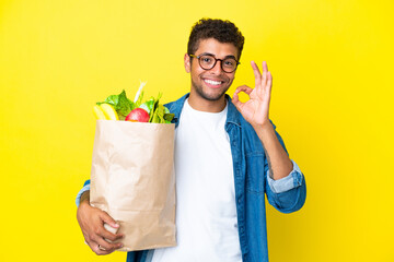 Young Brazilian man holding a grocery shopping bag isolated on yellow background showing ok sign with fingers