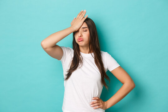 Young tired woman in white t-shirt, sighing and making facepalm to express annoyance, feeling fed up and tensed, standing over blue background