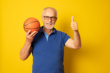 Naklejka premium Joyful senior man in casual polo shirt holding a ball and giving a thumb up isolated on yellow background