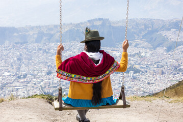 An indigenous Ecuadorian woman swinging on top of a tall mountain, looking down at the capital of Ecaudor, Quito. - obrazy, fototapety, plakaty