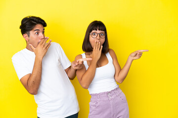 Young mixed race couple isolated on yellow background pointing finger to the side with a surprised face