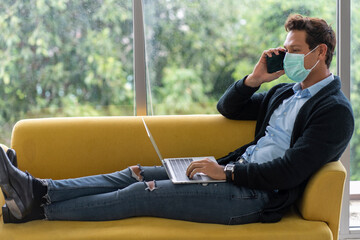Handsome male employee sits on a yellow sofa talking on the phone using a smartphone and is busy working on his laptop on his legs in the apartment living room. 
