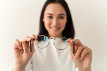 Positive smiling asian girl holding glasses and checking it cleanliness while posing on a white wall background. Focus at the hands with glasses