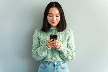 Indoor picture of adorable caucasian woman isolated on gray background smiling happily and being amused by content on screen of smartphone she is holding in both hands