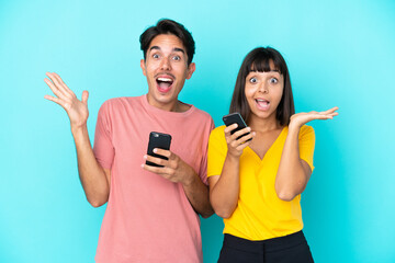 Young mixed race couple holding mobile phone isolated on blue background with surprise and shocked facial expression