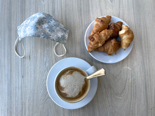 a protective mask on a breakfast table with coffee and croissants - coronavirus pandemic concept - new normal