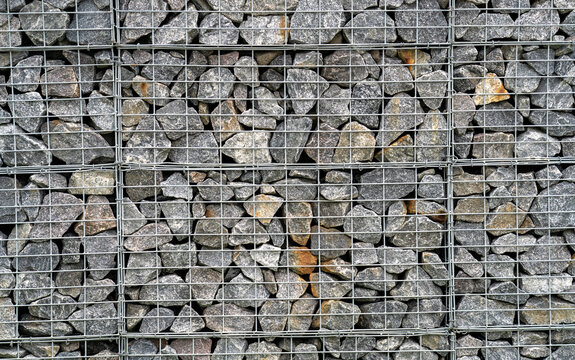 Wire Gabion Rock Fence. Stone wall texture, rocks behind metal grid fence background. Texture of fence made of many gray stones, reinforced with metal cage.