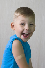 Naklejka premium A cute Boy in a blue T-shirt is laughing merrily, he has a patch on his shoulder, glued after the injection. The vaccine against Covid-19, vaccination of children. Healthcare. Positive emotions