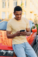 Vertical shot of the multiracial man sat on the bumper of his luxury red car and browsing internet at his smartphone with calm smile