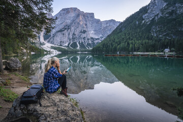 Woman reading e-book at Pragser Wildsee with reflected mountains on lake surface. Dolomites, Italy