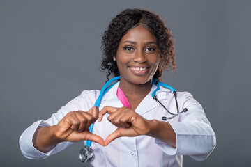 African woman doctor in a pink uniform, white coat and with a stethoscope holding a red heart....