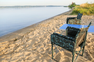 Camouflage fishing chairs and table on sandy beach near river, space for text