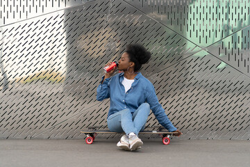 Tired african skateboarder girl refreshing after longboarding sit on skate drinking soda beverage...