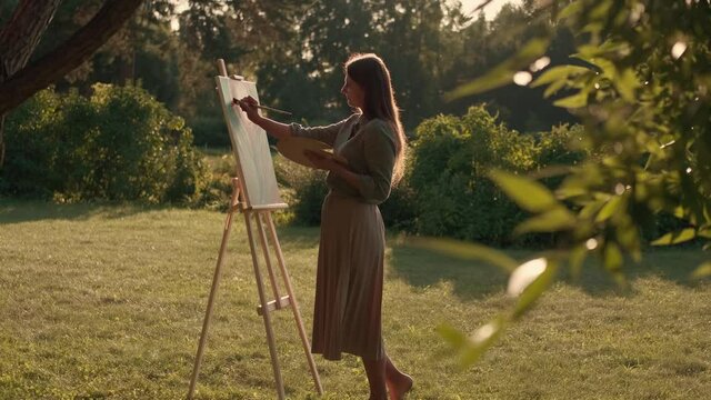 Creative woman artist paints a picture with a brush while standing near the easel in nature. Woman Artist, Art for Sales, Inspiration in Nature.
