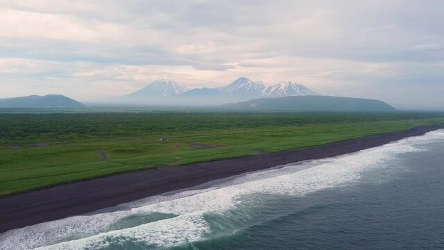Video from a drone of the volcanoes of Kamchatka, a summer evening view from the ocean with waves. Sunset with clouds and fog. Kozelsky, Koryaksky, Avachinsky.