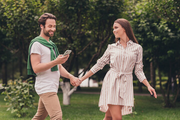 Fototapeta na wymiar Photo portriat young couple smiling walking in green park holding hands guy using smartphone looking on map