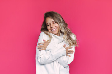 A young caucasian pretty smiling blonde woman with wavy hair in a white wool warm cashmere sweater enjoys the softness of the material hugging herself isolated on a bright color pink background