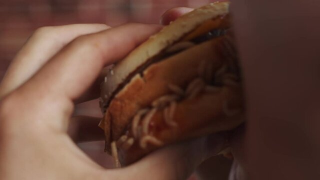 Man hand holds burger with maggots, bites and eats it, closeup. Man holds burger with maggots crawling on it and eats piece