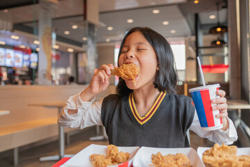 Close up portrait of a satisfied pretty  little asian girl eating fried chicken and french fries,...