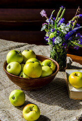 Ripe garden apples in a ceramic cup and a bouquet of wild flowers in a vase on a wooden table
